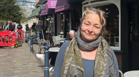 Holly Nichols/BBC  Kim Maclarty standing outside in a cobbled street. She has dark blonde hair with glasses perched on her head, a grey and green woven scarf around her neck and a grey long-sleeved top on. Behind her are shopfronts with tables and chairs in front and on the left a film crew is working between two red barriers. 