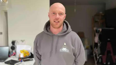 Joe Bishop, who served in the RAF, is pictured wearing a dark grey hoodie with the Building Bridges logo. He is a bald white man with hints of a ginger beard