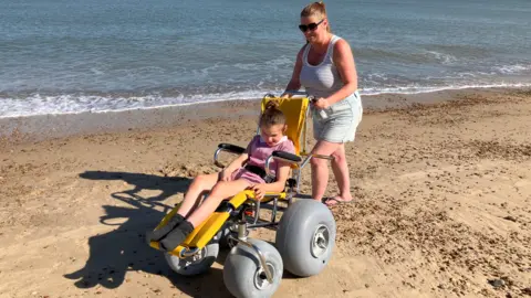 Jaimielee Rendall A woman wearing a vest and shorts pushing a beach wheelchair in which there is a young girl on sand.