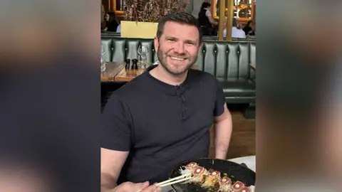 Gary Dawson A man smiles while holding chopsticks in front of a meal on a black dish on a restaurant table. He is wearing a black polo shirt. 