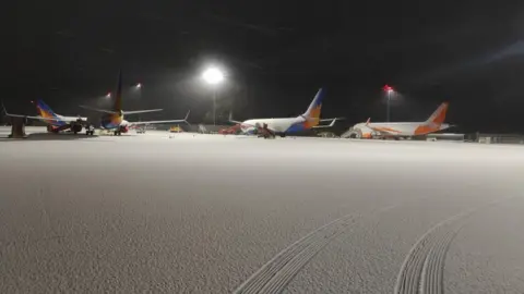 A night time view of Bristol Airport, with a powerful light showing three stationary aeroplanes and a covering of snow on the tarmac