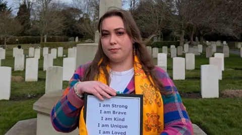 Claire wearing a colourful chequered jumper and a yellow scarf while standing in front of graves in a graveyard. She has long brown hair and is holding a piece of paper that says she is strong, brave, kind, unique and loved.