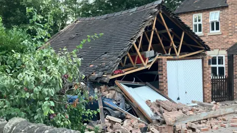 A double garage with its left door collapsed, bricks on the floor, and a car embedded in the garage after a crash