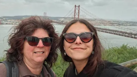 Claudia Andrade Claudia and her mum Idalina are wearing sunglasses posing for a selfie at a scenic viewpoint. A large red suspension bridge and a wide river with a cityscape are visible in the background.