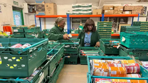 Two food bank workers, dressed in green fleeces, sort through donations. Green crates full of tinned food surround them on a wooden table, inside a warehouse.