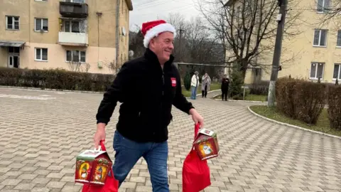 Tim Bamford A man in a black fleece and jeans, wearing a Santa hat, walks through a residential area carrying gifts.