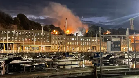 Image shows a large fire above a block of flats at night. There are also boats in the harbour in the foreground.