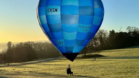 James Woodey A man sits in a small seat underneath a medium-sized hot air balloon close to the ground in the parkland of Ashton Court near Bristol. It is a sunny day, with the picture taken not long after dawn. The balloon is called a Cameron Superlight O-26 G-CKSW