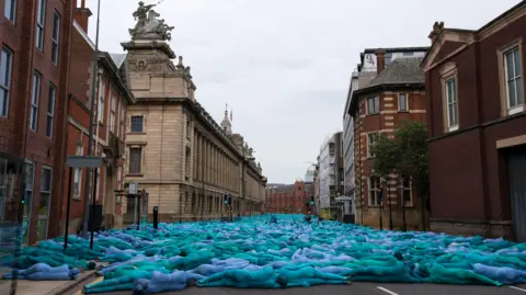 Getty Images/Jon Super Blue naked bodies facing away from the camera lie down across the road right up to the buildings on the left and right of the road. The image creates a blue river along the road.