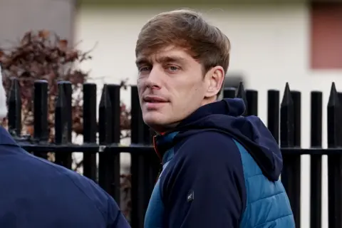 PA Media James Lawrence, who has light brown hair and wears a blue and navy coat. He has his back partially turned to the camera with his head tilted towards it.