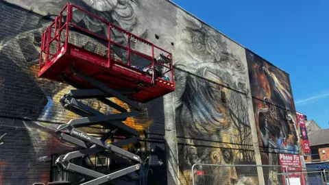 Alice Toon/ BBC A red cherry picker raised up to the mural, covering a third of it, the part of the mural that is visible shows a statue's face coloured in yellow and grey, on the right an image of an eagle with wings spread holding an oil lamp. There is a phone repair advertisement in the bottom right corner.