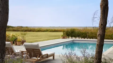 Omaze Two wooden sun loungers with beige cushions beside a swimming pool, with a lawn to one side and a view beyond of Blakeney Point 