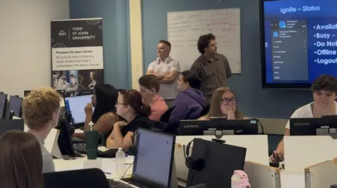 BBC / Samantha Whelan Staff and students sit in a room filled with desks. They have computers in front of them, and are looking to screens on the wall showing training materials. The walls are blue and white, with a whiteboard on the blue wall and a York St John poster leaning in the corner against where the white and blue walls meet.