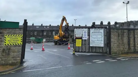 Julia Bryson/BBC A waste disposal site in Ilkley, West Yorkshire, showing a digger behind some gates, and a row of terraced houses in the background.
