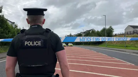 a police officer seen from behind, looking at a road where two police cars are visible and police tape reading "police line do not cross" has been strung upside down