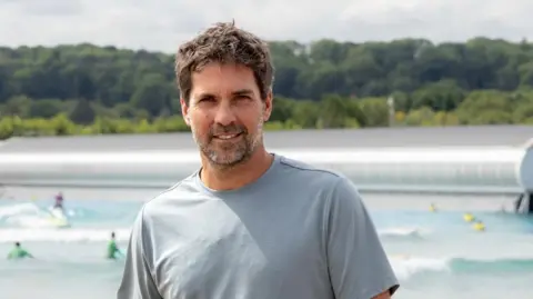 The Wave A man with dark ruffled hair and stubble wearing a grey shirt standing in front of a surfing lake with surfers on boards in the background and a forest of green trees behind the lake.