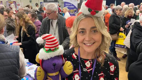 John Devine/BBC Hayley Mears has blonde, wavy, shoulder-length hair. She is wearing a Christmas top and a small Santa hat. She is holding a soft toy purple dog which has yellow paws and a yellow collar, and is wearing an elf hat.