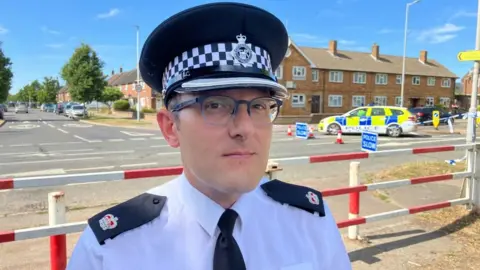 Sam Read/BBC Supt Lee Haines, in police uniform, looking at the camera by a police cordon in Luton, wearing a police hat and with glasses