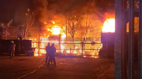 Two firefighters using a hose to tackle the fire which is behind fencing. The flames are high and orange and another firefighter can be seen to the side of the picture. 