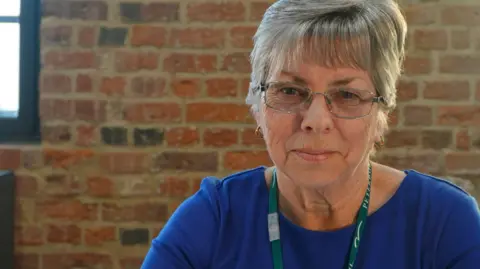 Peterborough City Council Geraldine stares at the camera. She wears a dark blue top and a green lanyard. Behind her is a brick wall with windows either side.