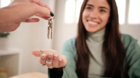 A smiling woman holds her palm out under a set of keys held by someone else out of shot. 