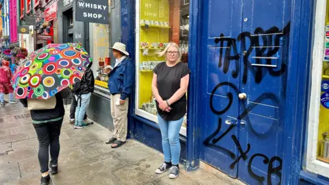 Jacqueline is standing beside her shop doors which are dark blue. They are covered in black graffiti. She is wearing a black t-shirt and blue jeans and has blonde hair and glasses. There is a passerby with a colourful umbrella.