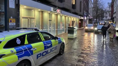 David Freezer/BBC Branch of HSBC bank with red paint splashed on the glass and doors. A police car is in the foreground. Other cars and people are on the paved street. 