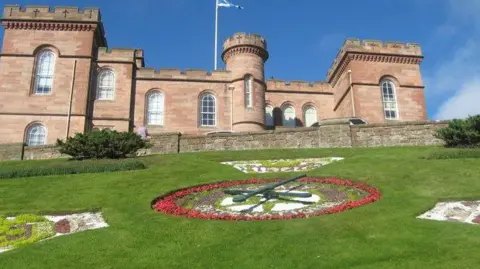 James Denham/Geograph The sandstone walls and faux battlements of Inverness Castle above a grassy slope with flower beds and the floral clock.