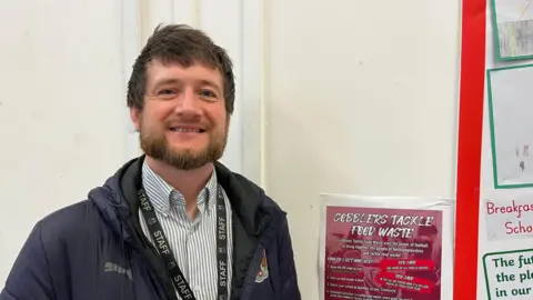 A man in a Northampton Town branded jacket stands next to a poster promoting food waste reduction 