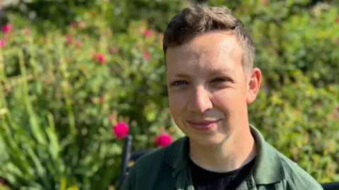 A young man, aged 30 with short brown hair looks into the camera with blurred pink roses behind in a garden.