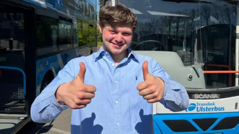 Luke Wilson puts two thumbs up as he stands in front of two, parked Ulsterbuses. He is a young man with short, light coloured hair and a blue shirt. 