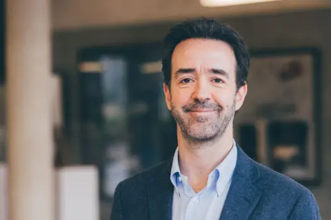 University of Essex A man with short dark hair, stubble and a blue shirt and blazer smiles at the camera.