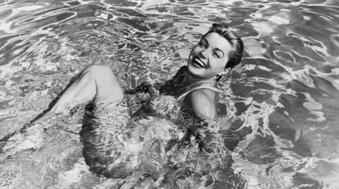 Getty Images Esther Williams in a swimming pool. She is on her back with her knees bent and head turned towards the camera, smiling. 
