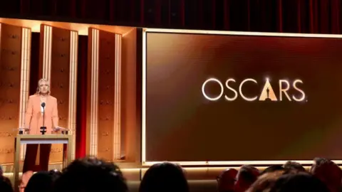 REU A woman in a salmon coloured suit stands on a large stage next to a sign reading: 'OSCARS'