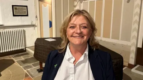 Ben Parker/BBC A woman is standing in a room which looks like an old town hall. She has medium length blonde hair and is smiling. She is waring a blue jacket and white top. 