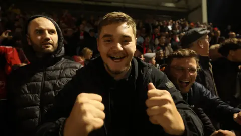 PA Media Three male Kettering Town fans in the crowd, all dressed in black puffer jackets, look jubilant at their side wins over Northampton Town. The central man has both his fists raised in triumph. 