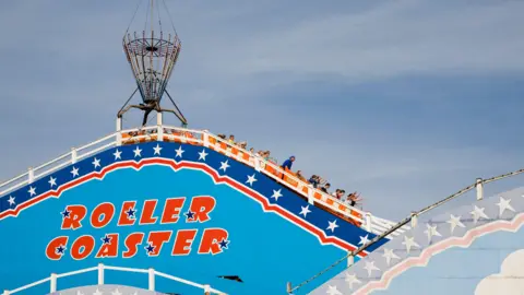 People in carriages heading downwards. Some in the front carriages have their arms in the air. American-style red writing saying "roller coaster" is painted on a sky-blue side of the ride with white stars, and against a navy and red trim bordering the ride. The carriages are orange.