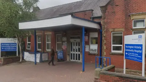 Google A street view image of Accrington Victoria Hospital, a red brick building with blue and white signs either side of a white awning with blue legs over the entrance and a man walking under it to the front door