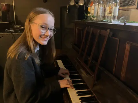 BBC/Amy Holmes An image of a teenage girl playing a piano. She is smiling and is wearing glasses and has shoulder length brown hair.