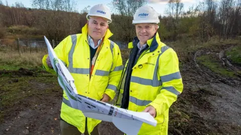 Arthur Mann and Andrew McArthur standing outdoors in land - they are both wearing high vis jackets and hard hats, while holding plans showing a proposed new housing development