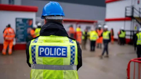 Image of the back of a police officer, wearing a yellow florescent jacket and a blue hard hat. In front of him, blurred out a focus, people can be seen wearing the same jackets and some in full orange florescent work-wear.