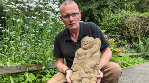Vindolanda Charitable Trust Dr Andrew Birley, a middle-aged man with a shaved head and glasses - wearing a black polo shirt and brown trousers - holding the sculpture. It is a warm beige rock in the shape of a humanoid figure with pupil-less eyes. It holds a long curving object in one hand and a circular object in the other. Although the edges of its body seem to have become rough and worn-down, the face of the figure is instantly identifiable.