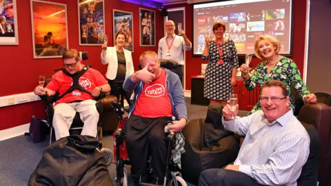 Service users at The White Lodge Centre in Chertsey, Surrey, alongside employees of the company pictured after a cinema room was installed. Staff are all holding up a glass of fizz and there are two service users in wheelchairs.