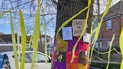 Empics Campaigners sitting and standing next to the oak tree, which has been covered in signs and yellow ribbons. There are cars parked behind it, and further back is a metal fence and modern homes. There is a police car and a police officer in the background.