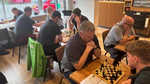 Simon Thake/BBC A group of men play chess on small square tables in a cafe.