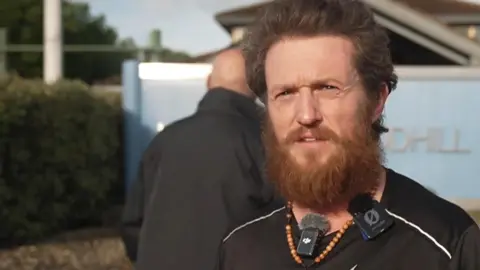 Tommy Robinson is wearing a black t-shirt and has two microphones attached to the collar. He is also wearing rosary beads around his neck attached to a wooden crucifix. Behind him are two men and a large blue sign for HMP Woodhill. Tommy has a long beard and hair.