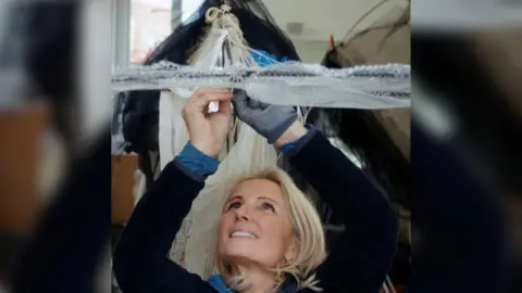 Tessa Campbell Fraser The artist Tessa Campbell Fraser, a middle-aged blonde woman, wearing a dark top and reaching above her head as she works on one of her pieces about whales.