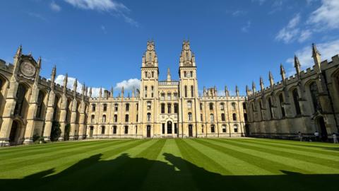 The sun shines on an Oxford college sandstone building surrounding a grass quadrangle with blue skies behind