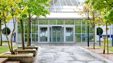 File image from 2015 showing the walkway and entrance at Woolwich Crown Court. The building has a glass facade with three glass doors in the centre - the centre is a revolving door. The path towards the court is lined with small trees.