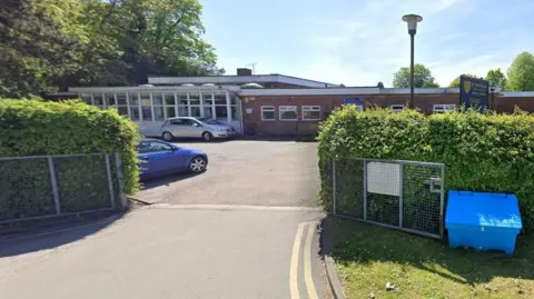 Google School entrance with hedges either side and a road leading into a car park where a blue and a silver car are parked. There is a single-storey school building beyond with a white block to the right, with a facade of mainly windows, and a brick building to the right, with smaller windows.